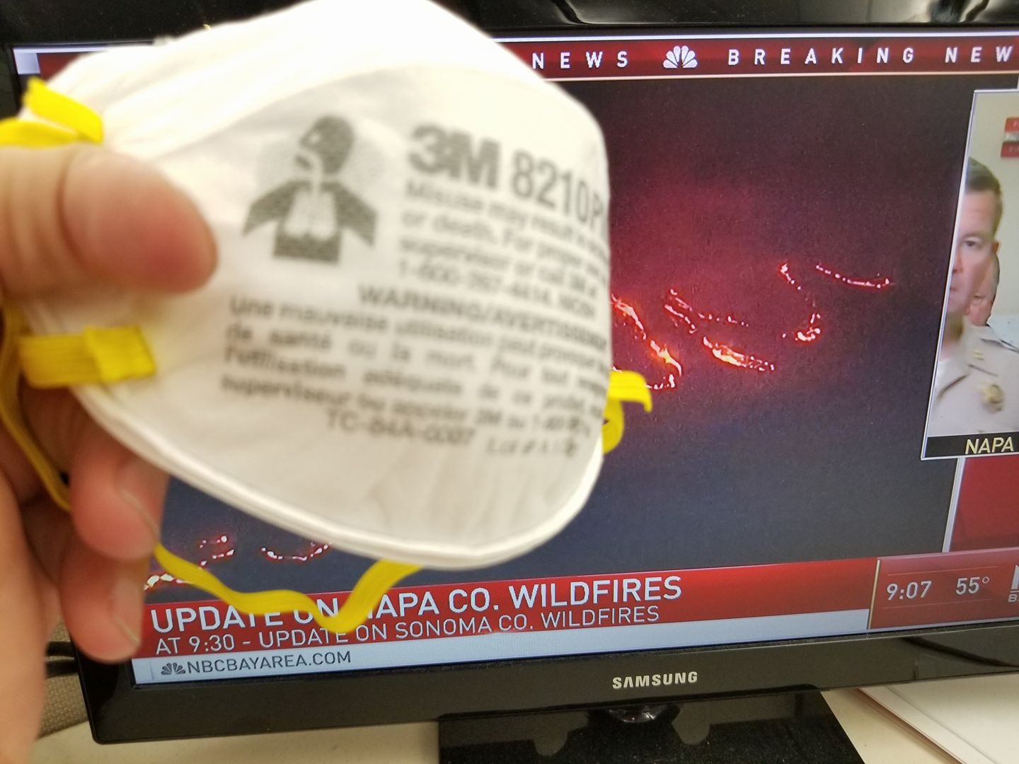 An N95 mask held up against wildfire coverage on screen